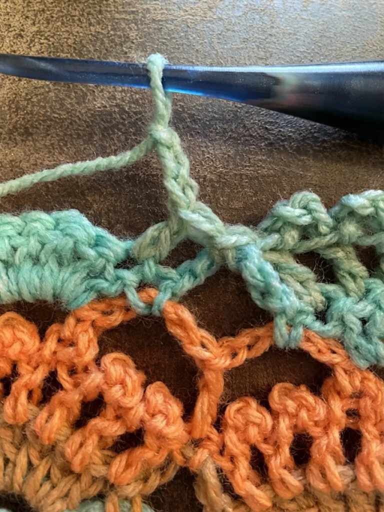 Close-up of a blue crochet hook pulling up a light green chain loop to form a picot above rows of orange and green crochet fabric on a brown leather surface.