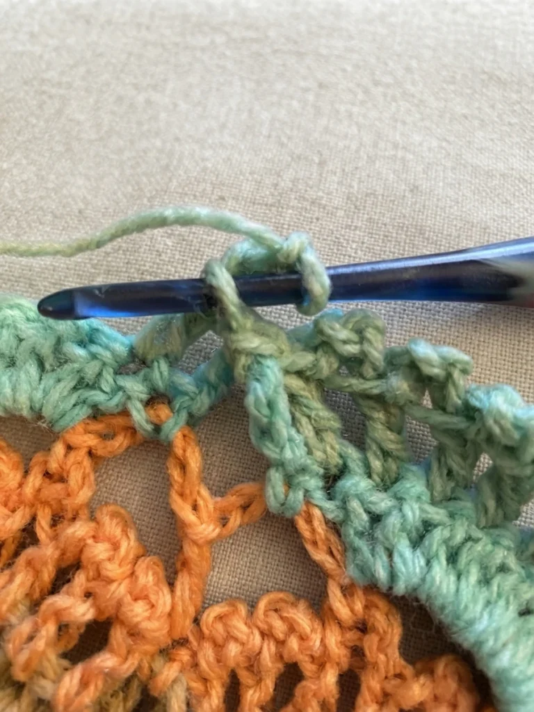Close-up of a dark blue crochet hook holding a loop of sage green yarn while working a picot stitch, with a green and orange crocheted swatch resting on a light beige fabric surface.