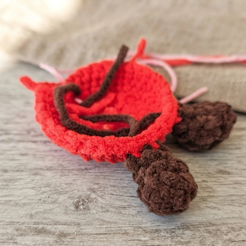 Work in progress amigurumi apple: a partly crocheted red body shaped like a small bowl with two brown crocheted legs attached at the bottom, loose pink and brown yarn ends and a brown stem piece resting inside, placed on a light wooden surf