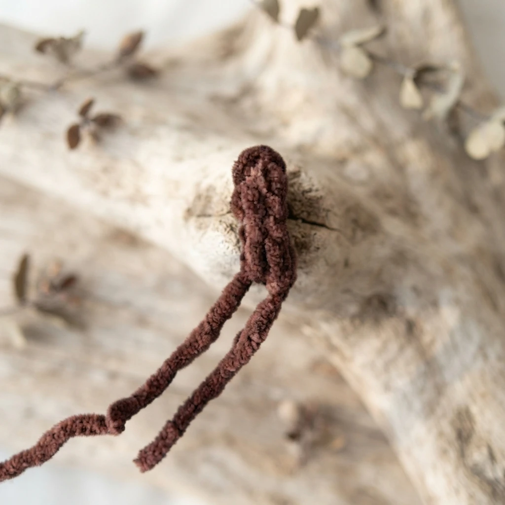 Small brown crocheted stem piece with two long yarn tails resting on a pale weathered branch, with blurred dried leaves in the background