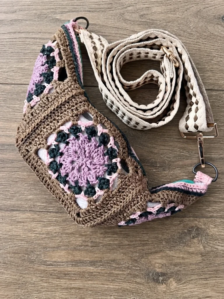 Crocheted crossbody bag with a granny square motif in brown, purple, pink and dark green, with a coiled woven strap on a wooden surface.