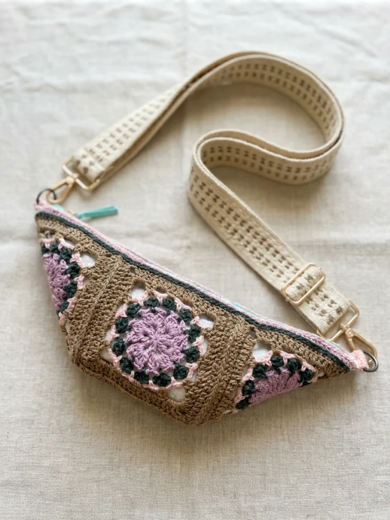 Crochet crossbody belt bag with granny square motifs in mauve, dark green and tan, finished with a cream woven adjustable strap and mint zipper, shown lying flat on light linen fabric.