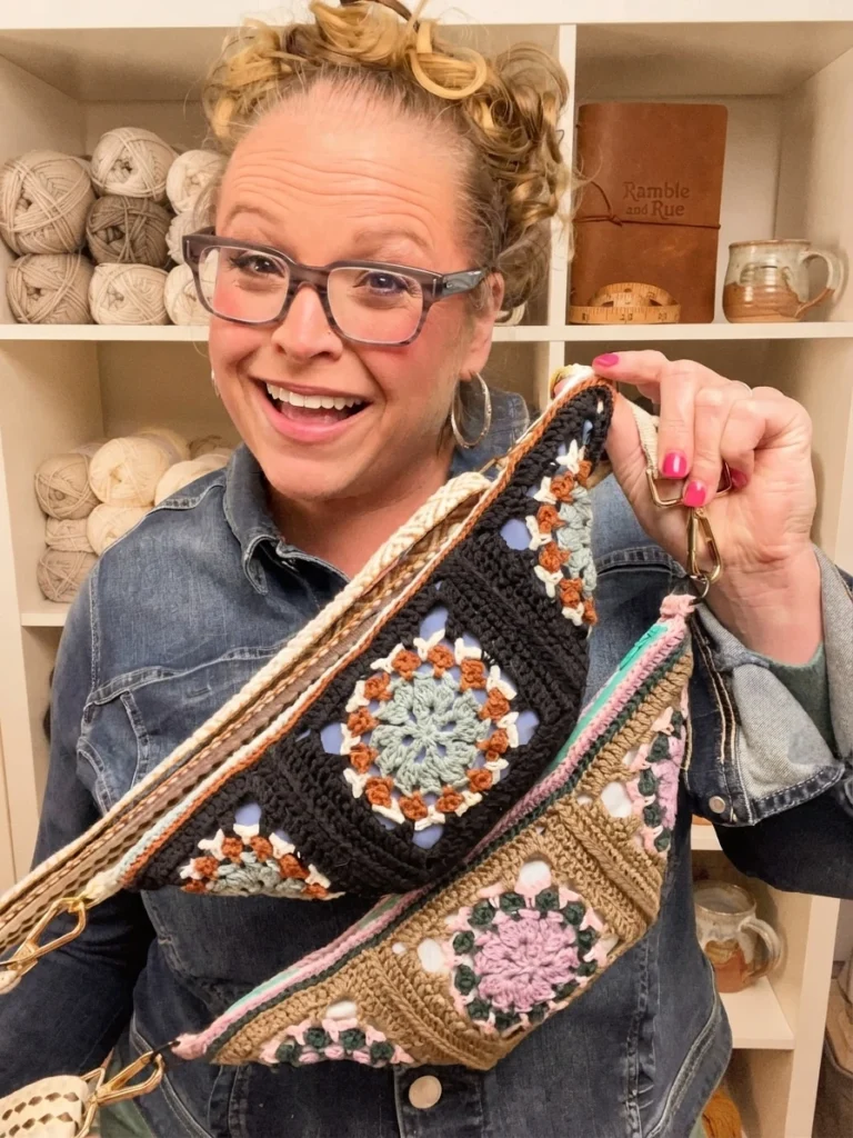 Smiling woman in denim jacket holding two crocheted crossbody bags with granny square motifs in front of yarn shelves