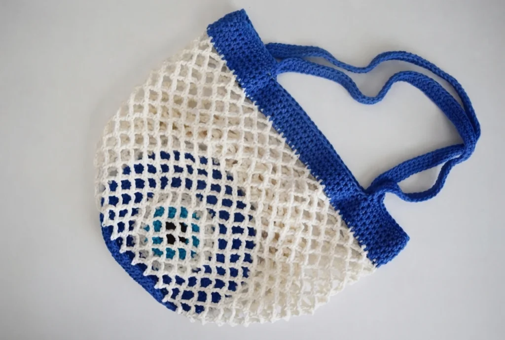 Crocheted market bag with white open mesh body, blue top band and handles, and a concentric blue, white and turquoise eye motif visible through the mesh on a light background
