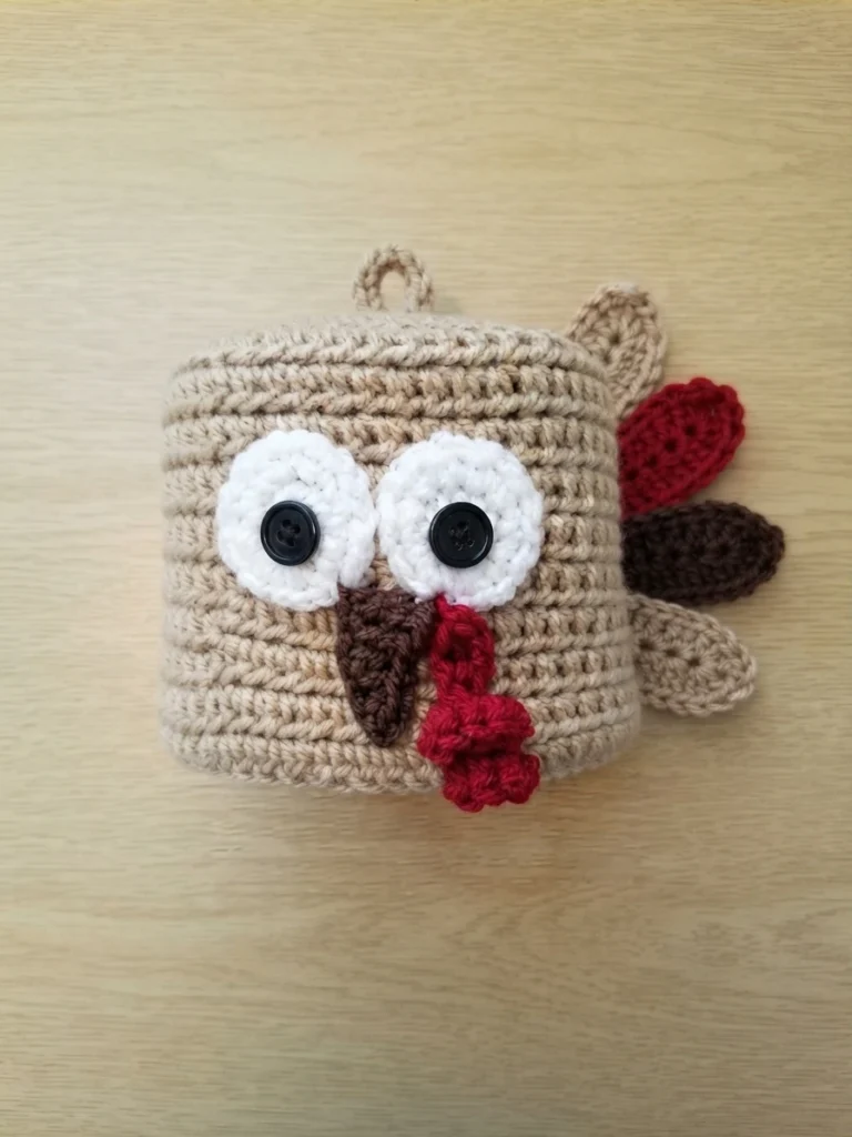 Crocheted tan turkey toilet paper roll cover lying flat on a light wood surface, with two round white eyes, black buttons, a brown beak, a red snood and red, brown and tan feather shapes along one side.
