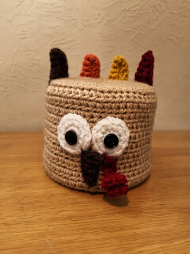 Crocheted tan turkey toilet paper roll cover with two round white eyes, black button pupils, a brown beak, a red wattle and four upright colored tail feathers, standing on a wooden surface.
