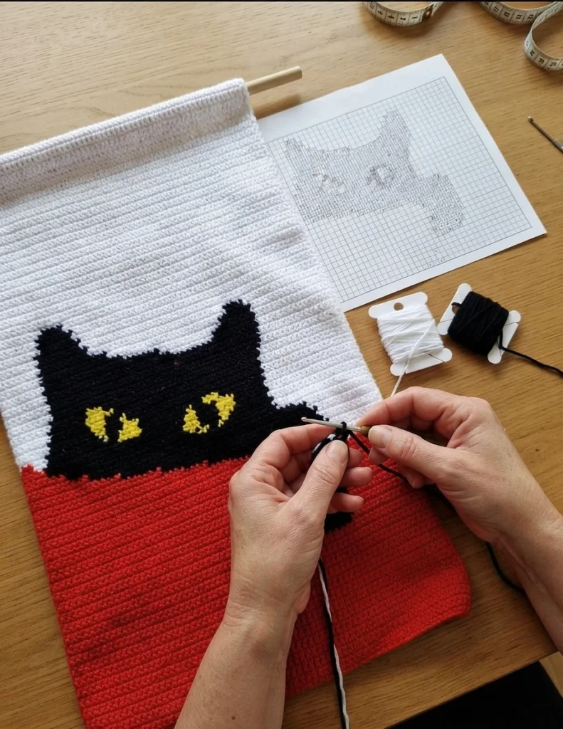 Sac tote crocheté rouge et blanc avec un chat noir aux yeux jaunes, posé devant un buisson de roses jaunes, puis en cours de réalisation sur une table en bois avec un graphique papier et des pelotes de fil.