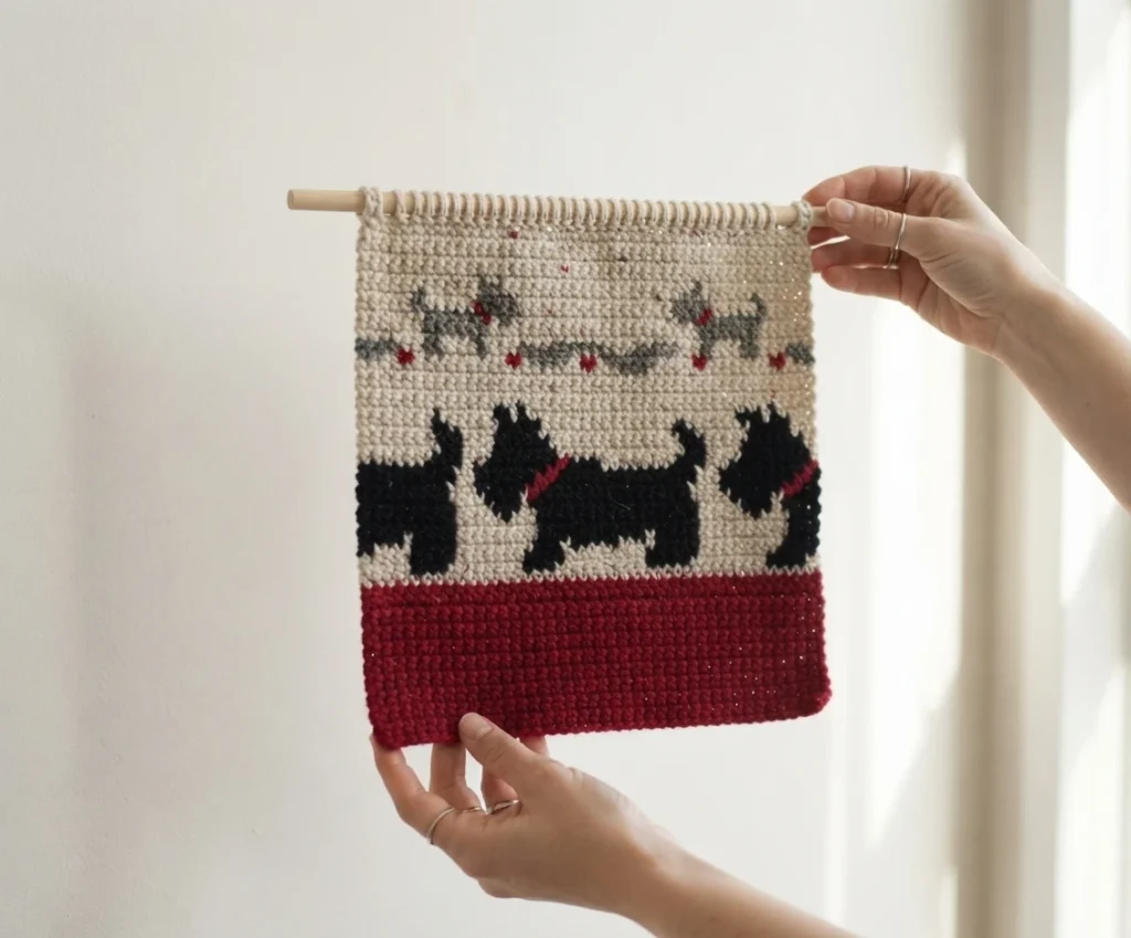 Hands holding a cream crochet tapestry on a wooden dowel, showing black dog motifs and a red band along the bottom edge.