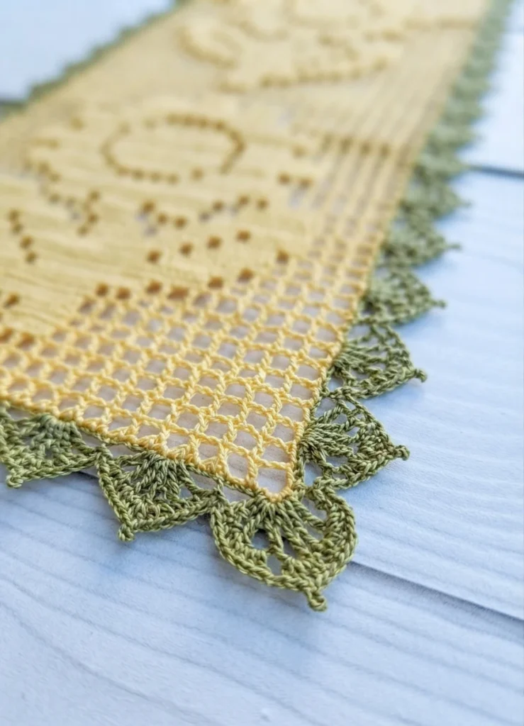 Yellow filet crochet table runner with an open mesh sunflower motif, edged in a green scalloped lace border, shown at an angle on white painted wood.