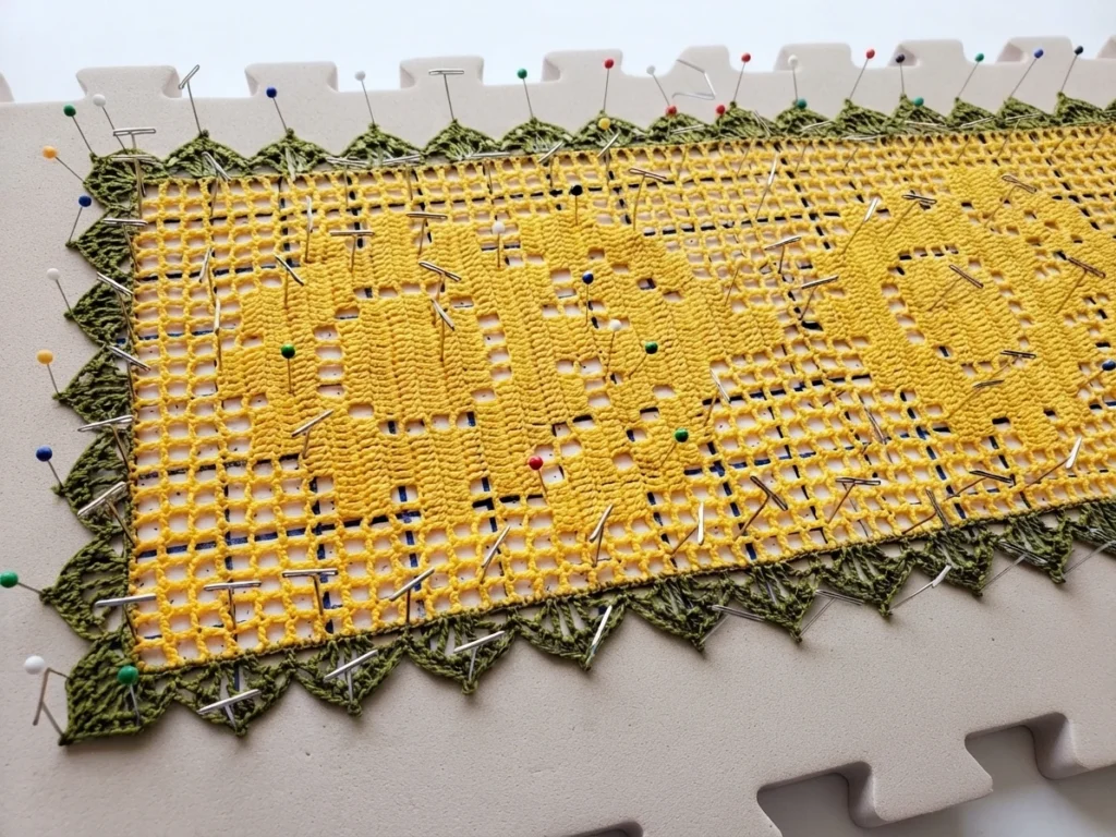 Yellow filet crochet table runner with a green scalloped border stretched flat and held with pins on a blocking mat, the sunflower motif showing through the open mesh