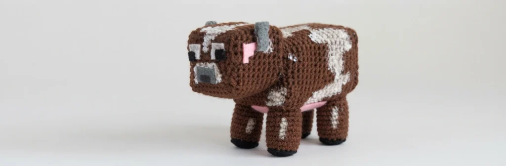 Brown and white crochet blocky cow with grey felt horns, pink felt ears, square eyes and pink udder