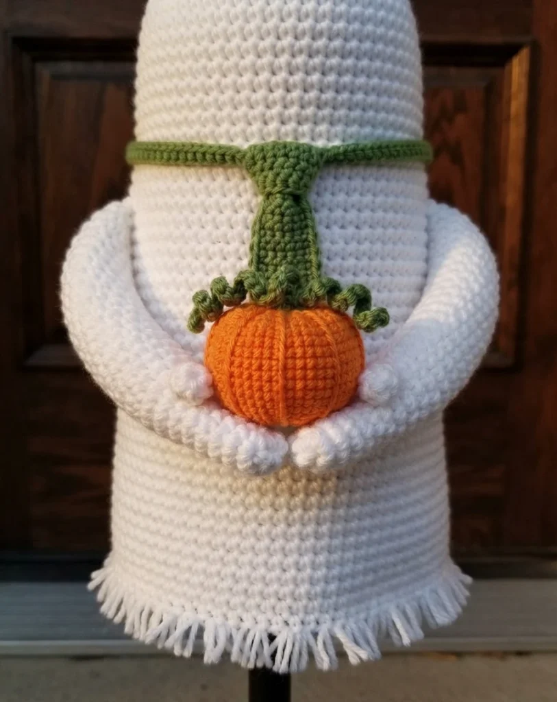 Close-up of a white crocheted ghost wall hanging with black button eyes and a stitched smile, a green crocheted neck band and scarf tail, and white fringe along the bottom edge, its arms wrapped around an orange crocheted pumpkin with a gre