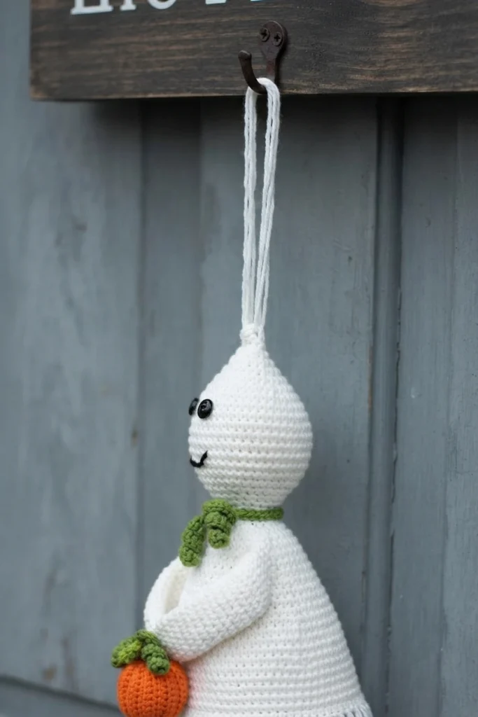 Crocheted white ghost with black button eyes and a stitched smile, wearing a green scarf and holding a small orange crocheted pumpkin with green leaves, hanging by a white cord from a metal hook on a wooden sign against grey planks.