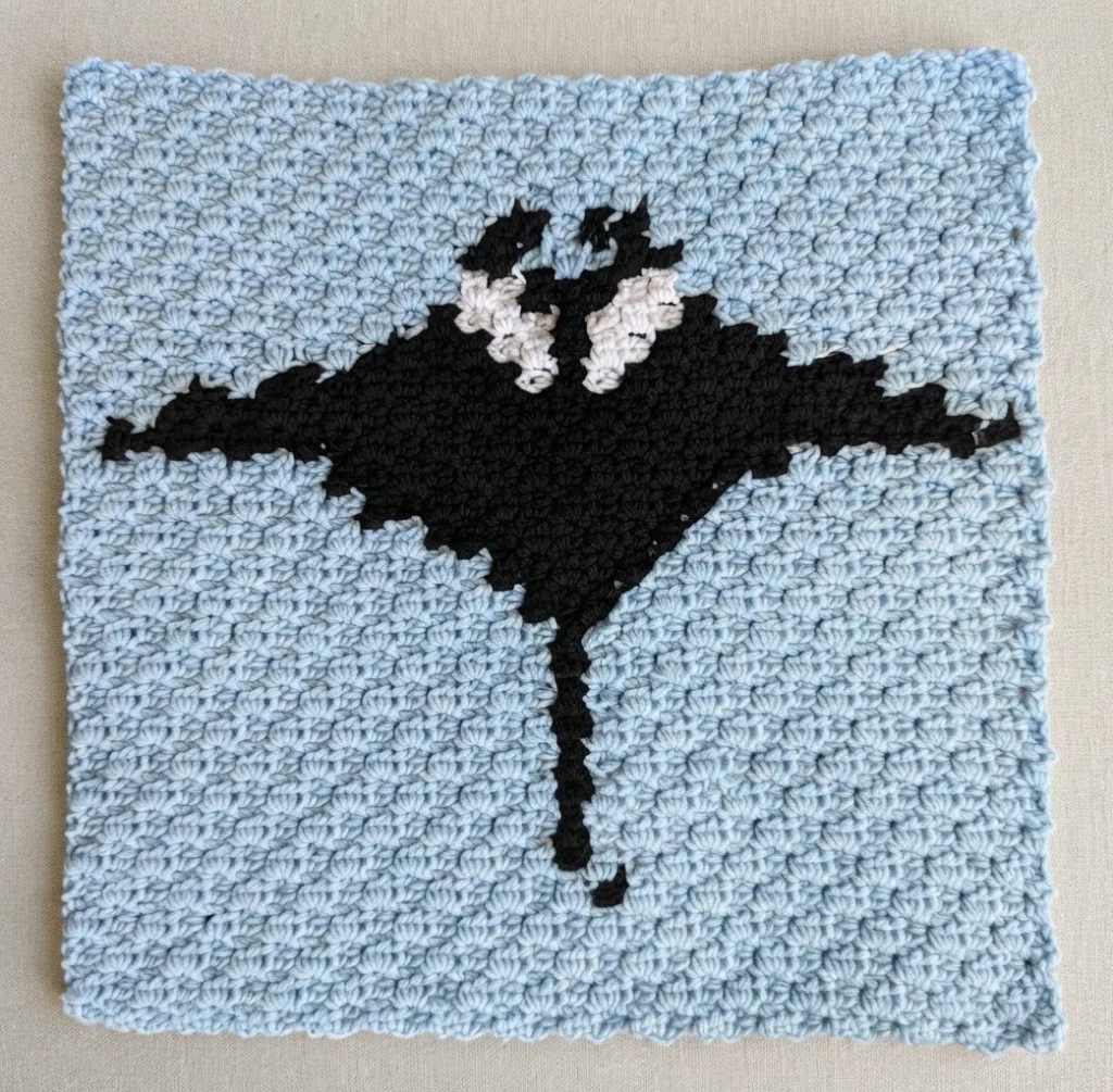 Light blue crocheted square block with a black manta ray motif and white markings, on a light beige background.