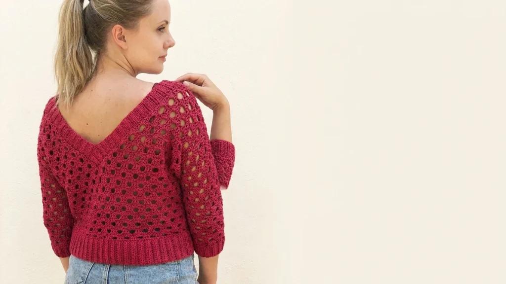 Woman seen from behind wearing a red openwork crochet sweater with a V-shaped back neckline, ribbed hem and three-quarter sleeves, paired with blue jeans against a light wall.