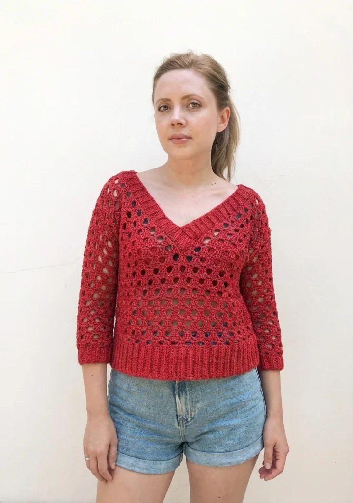 Woman wearing a red crocheted V-neck sweater with openwork pattern and denim shorts against a plain light wall