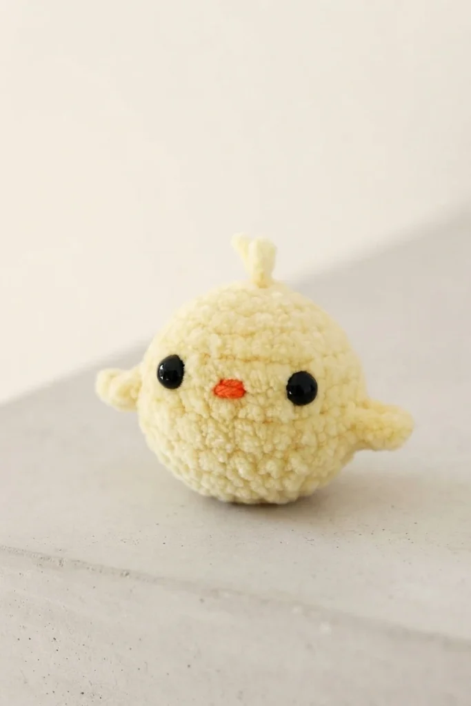 Single pale yellow crochet chick with black eyes, orange beak, small wings and a tiny head tuft, sitting on a light surface.