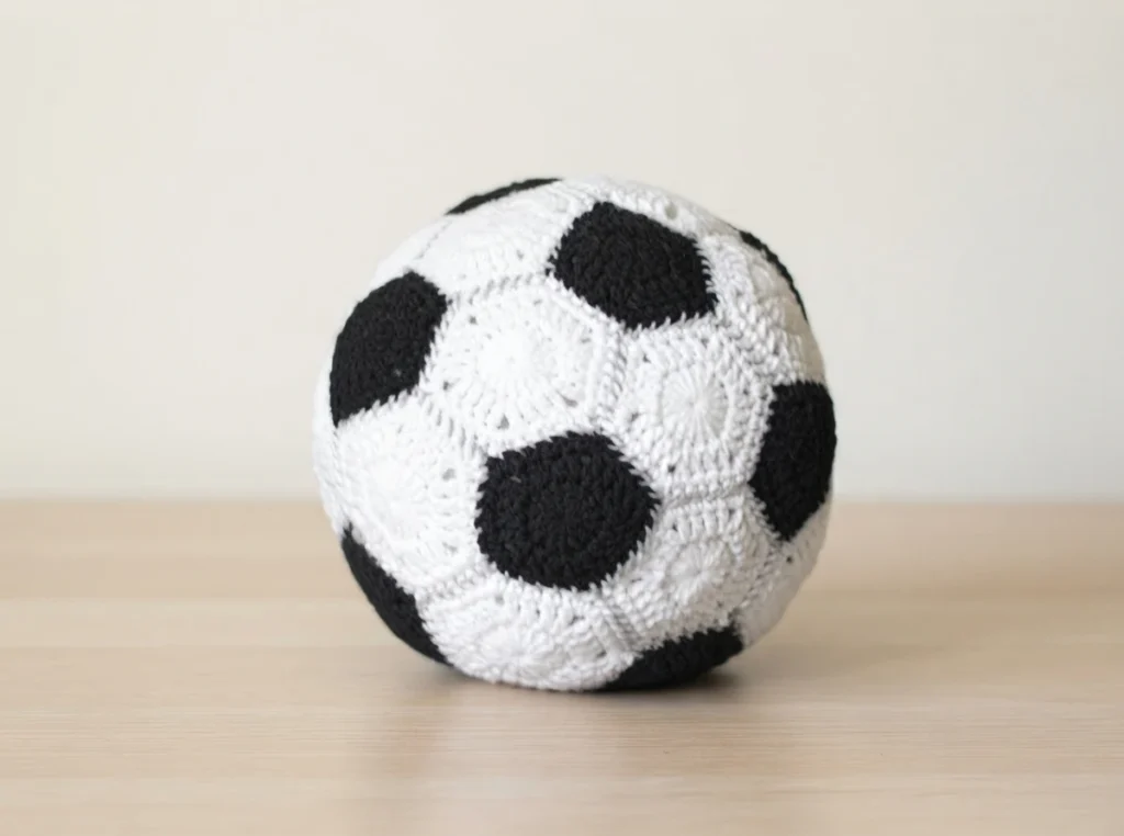 A crocheted black-and-white soccer ball made of pentagon and hexagon motifs, resting on a light wooden table against a plain cream background.