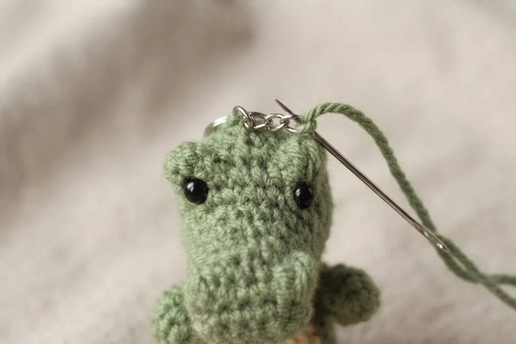 Close-up of a green crocheted alligator head with a silver keyring chain being whipstitched to it with a tapestry needle threaded with green yarn.