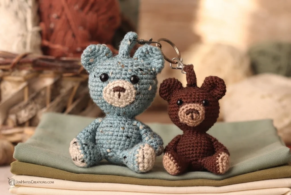 Two small crochet bear keychains, one speckled blue and one brown, sitting on folded fabric with yarn in the background.