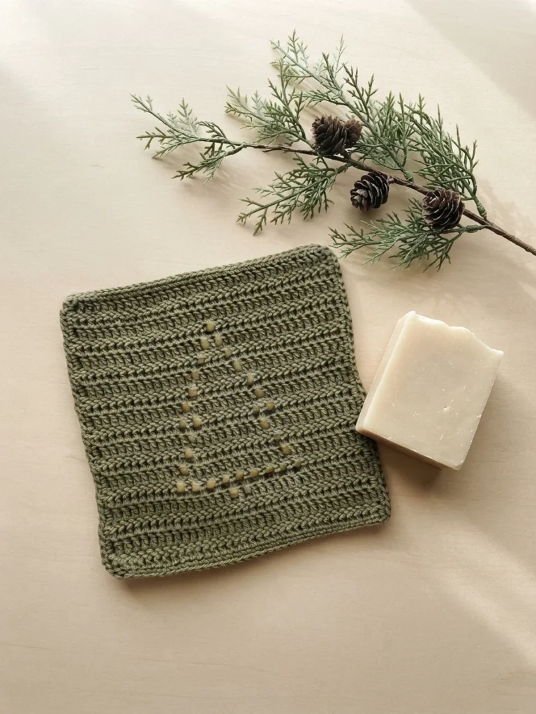 Olive green crocheted washcloth with an openwork fir tree motif, placed beside a white soap bar and an evergreen sprig with pinecones.