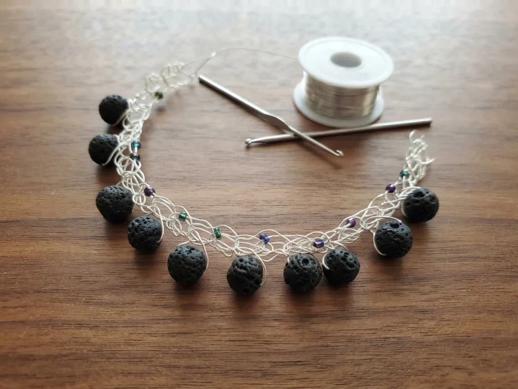 Wire-crocheted silver bracelet with black lava rock beads and small iridescent seed beads, photographed on a wooden table next to a spool of silver wire and two crochet hooks.