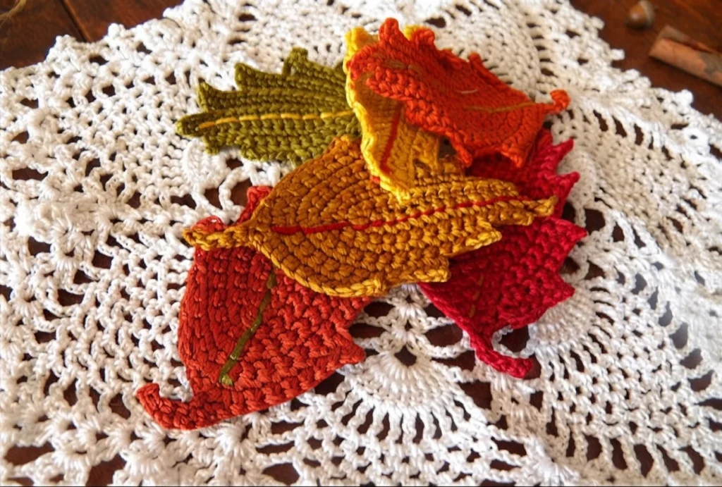 Un amas de feuilles crochetées rouges, orange, jaunes et vert olive repose sur un napperon blanc ajouré.