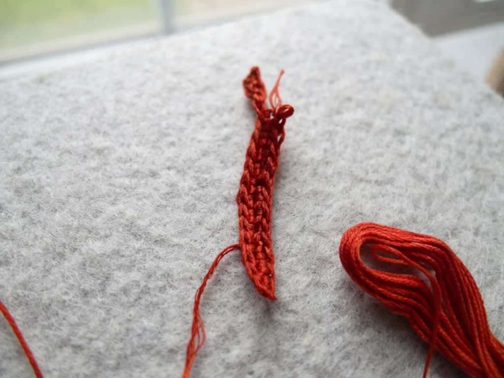 Pièce crochetée rouge étroite, avec des fils lâches et une réserve de fil posées sur un tissu clair.