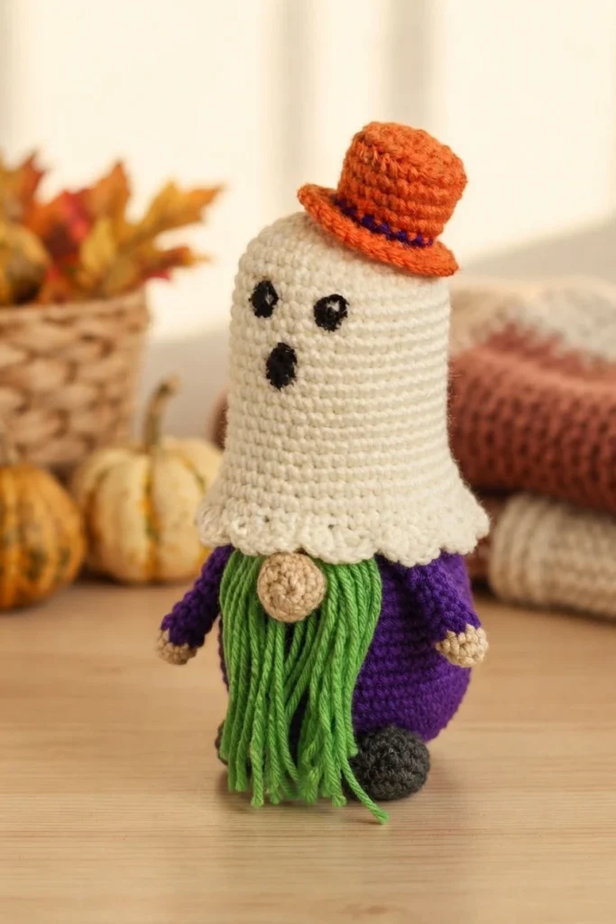 Crochet ghost gnome with a white hooded ghost head, black embroidered eyes and mouth, a small orange top hat with a purple band, a green yarn beard, a beige nose and a purple body, standing on a wooden surface with autumn leaves, pumpkins,