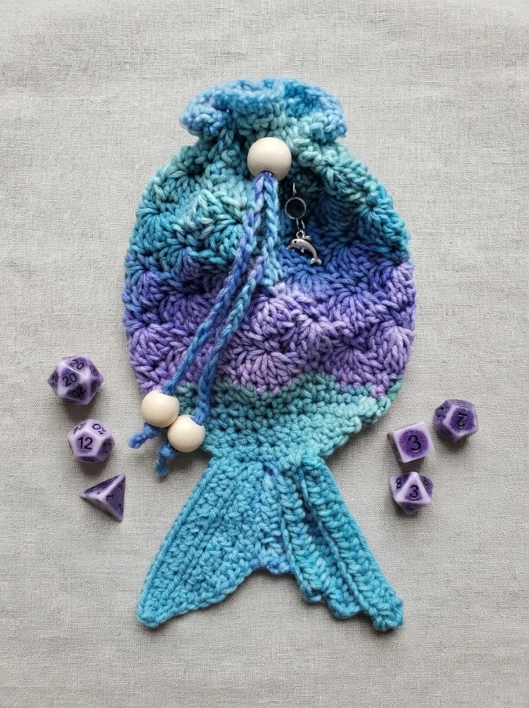 Crocheted mermaid tail dice bag in blue and purple gradient yarn with a drawstring closure, wooden beads and a small dolphin charm, surrounded by purple polyhedral dice on a light fabric background.