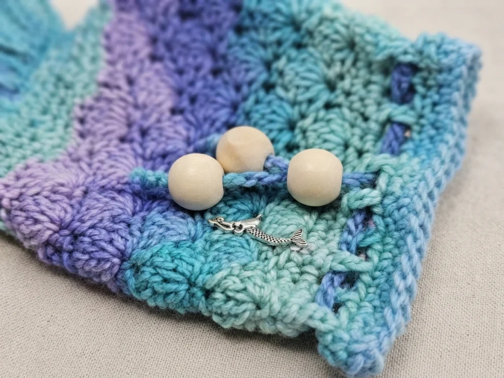 Close-up of a crocheted mermaid tail dice bag in blue, teal and purple with three wooden beads on the drawstring and a small mermaid charm