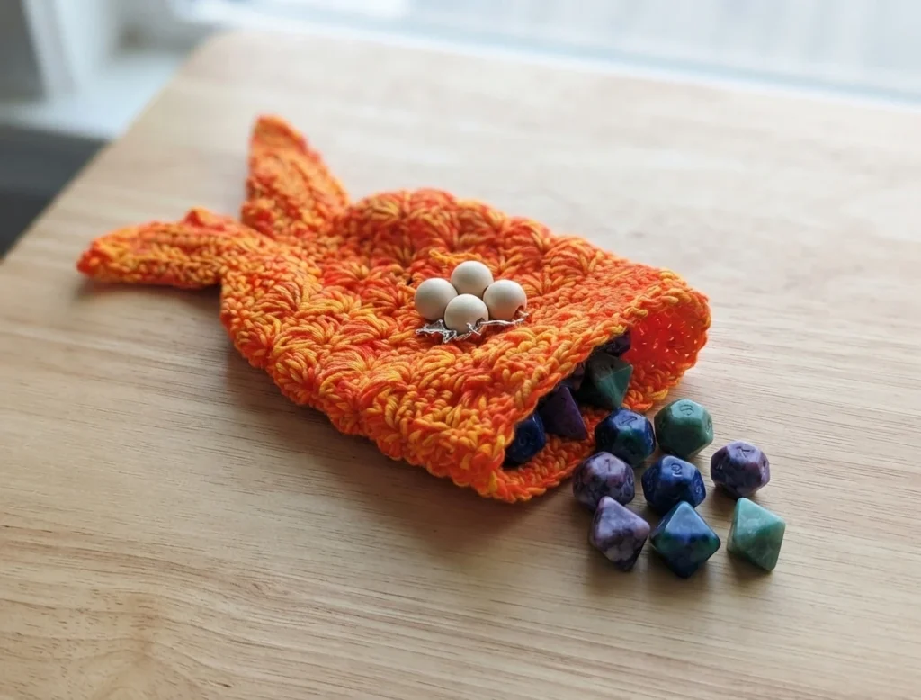 Orange and yellow crocheted mermaid tail dice bag with wooden bead drawstring and starfish charm, shown open with multicolored dice spilling out on a wooden table