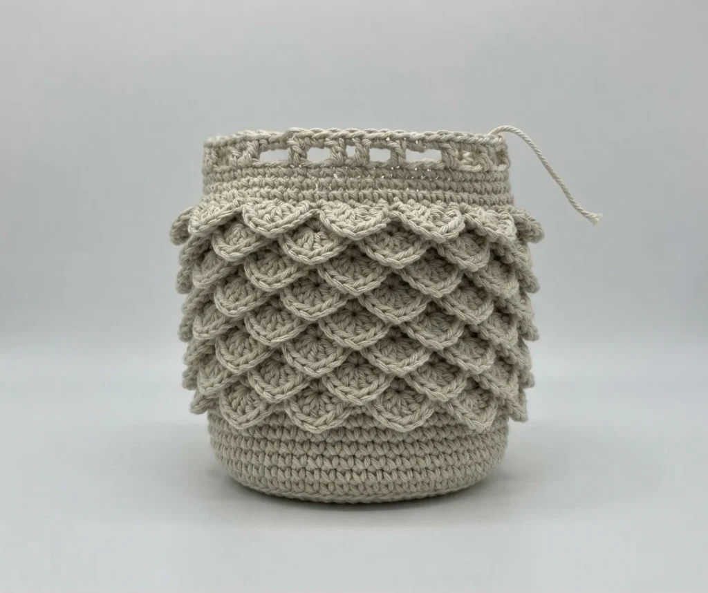 Beige crochet bag with overlapping crocodile stitch scales, open top and loose yarn tail, on a plain light background