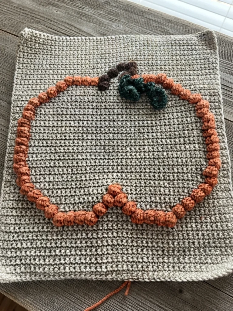 Flat crochet panel in light gray yarn with an orange bobble stitch pumpkin outline, brown bobbles forming the stem and dark green bobbles forming a leaf, laid on a wooden surface with a loose yarn end at the bottom.