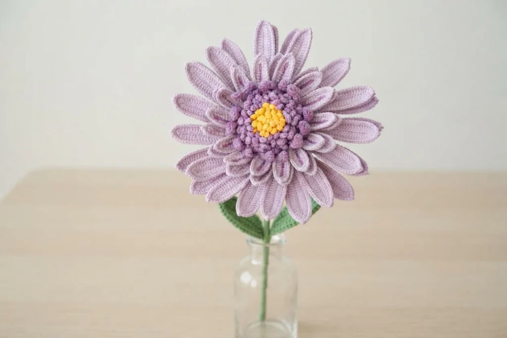 Crocheted lilac gerbera daisy with a purple textured center and yellow middle, standing in a clear glass vase on a light wooden table