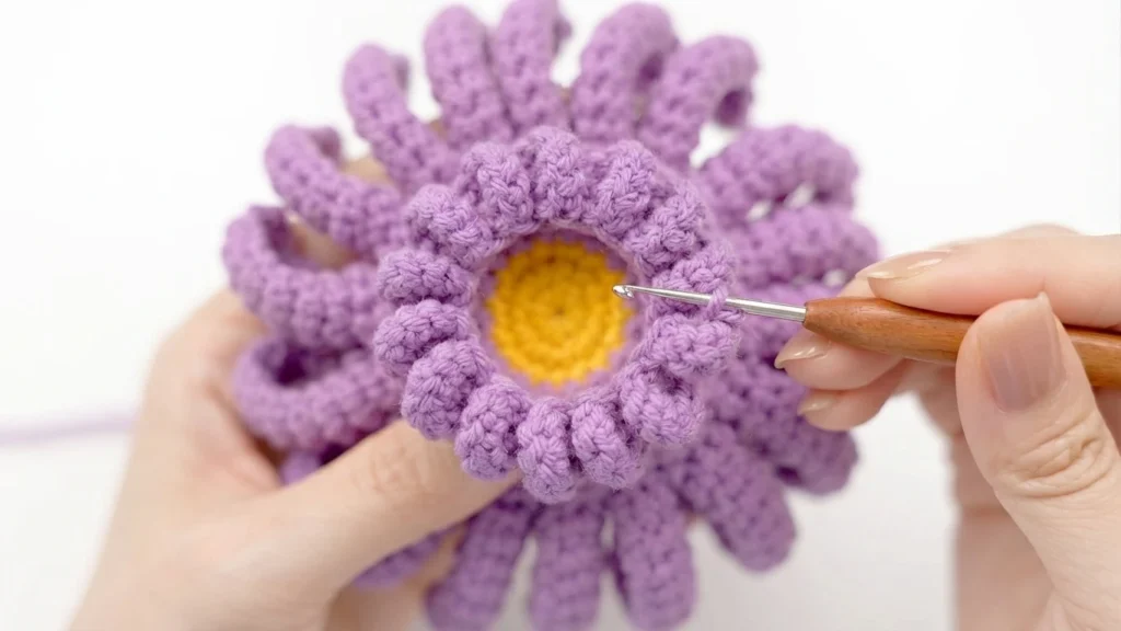 Close-up of hands holding a crocheted flower center with small purple petal loops around a yellow middle, with a wooden crochet hook on a white background