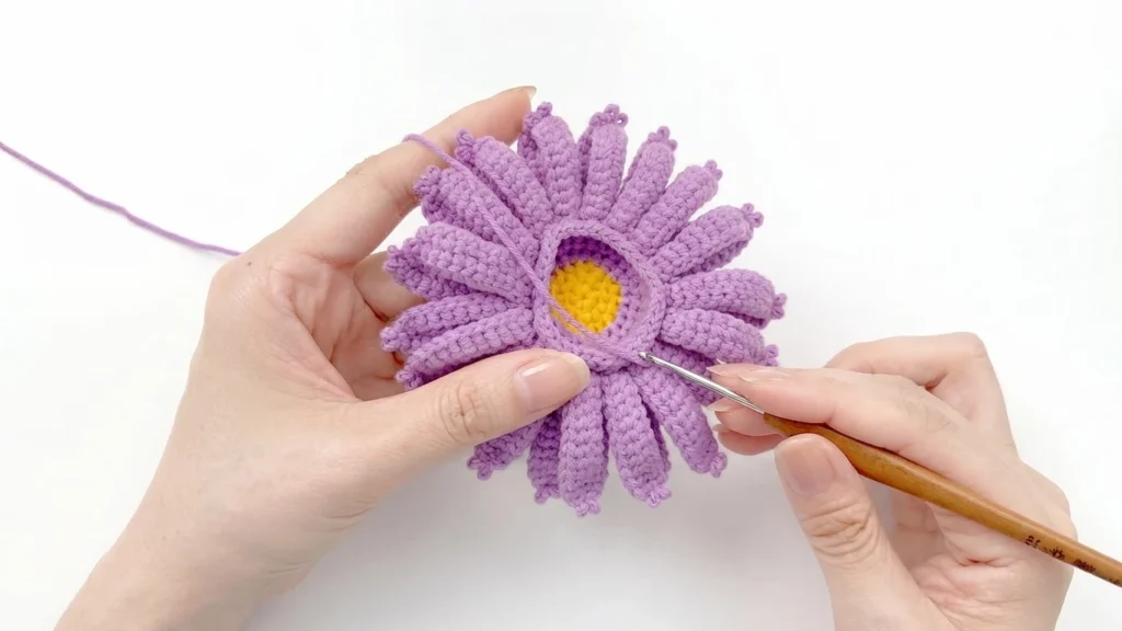 Hands crocheting purple petals around a yellow center with a wooden handled hook on a white background