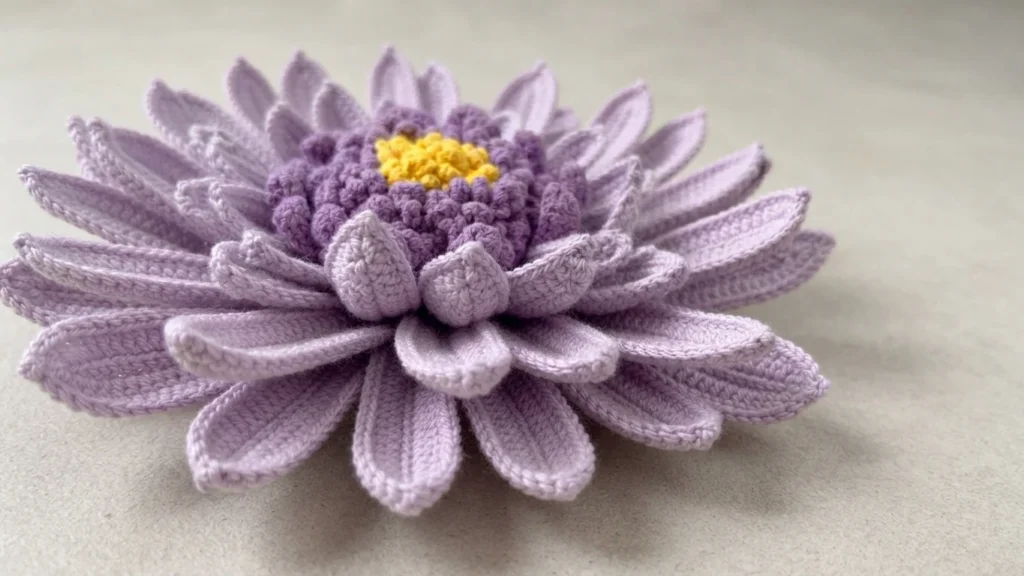Crocheted light purple gerbera daisy with layered petals and textured purple and yellow center on a light surface
