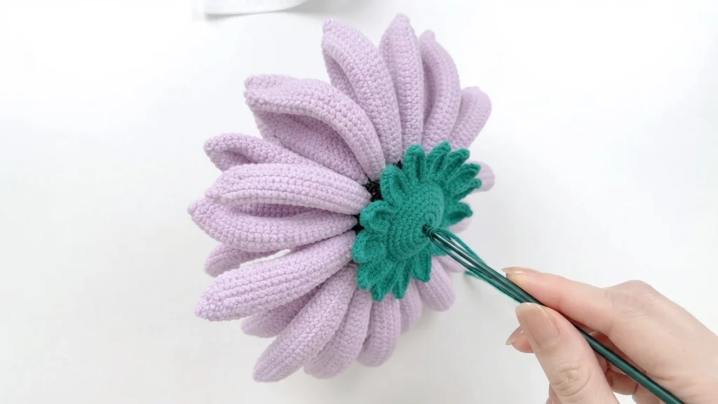 Hand inserting a dark green stem into the green crocheted base on the back of a lavender gerbera flower