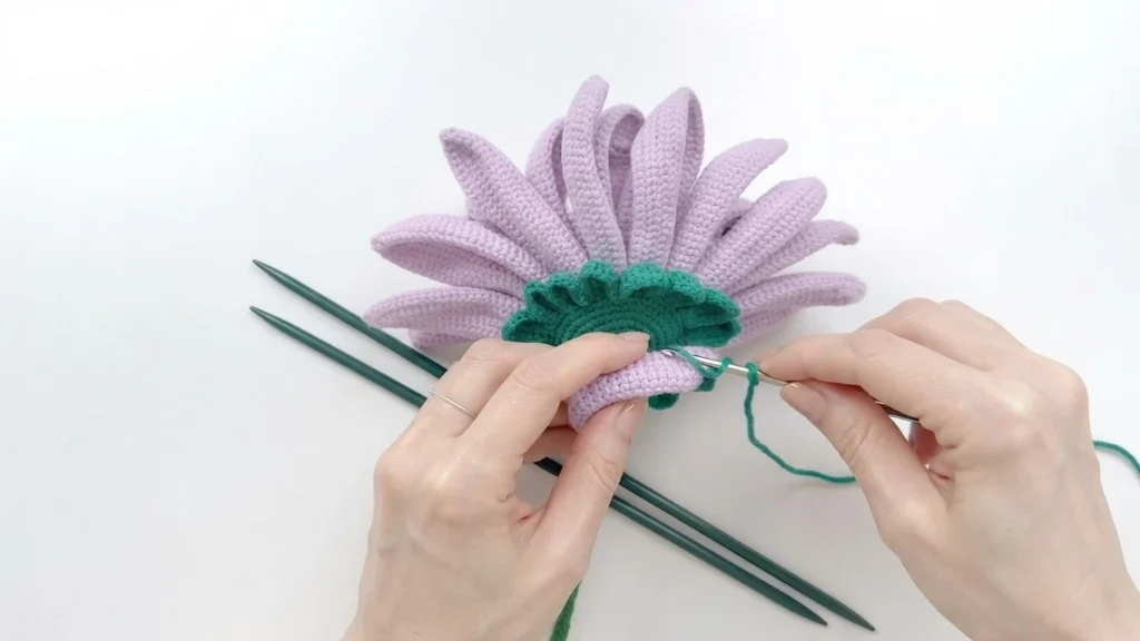 Hands sewing a green crocheted base onto the back of a light purple crocheted gerbera flower, with long green stems beside it on a white surface.