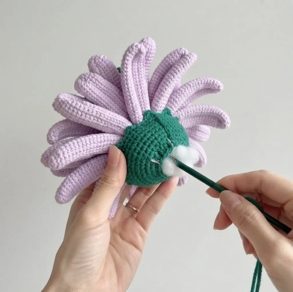 Hands pushing white fiberfill into the green base of a purple crochet gerbera daisy with a dark green tool.