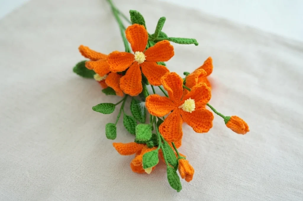 Orange crochet hibiscus flowers with pale yellow centers, green stems and leaves, and two small buds resting on light fabric.