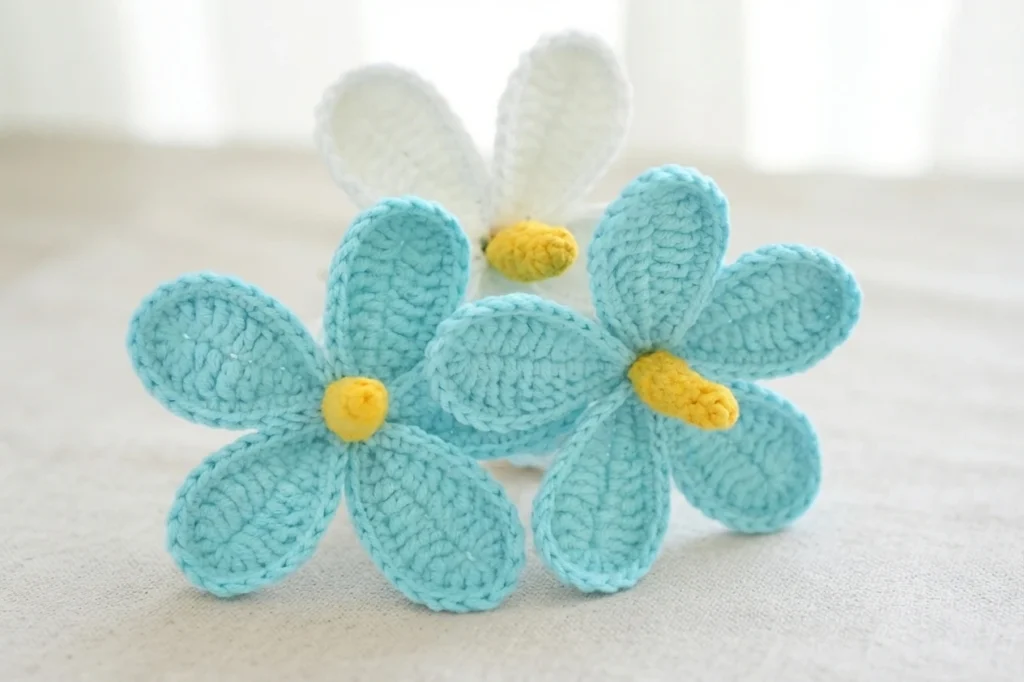 Three small crocheted five-petal flowers, two turquoise and one white, each with a yellow center, arranged on a beige fabric background.