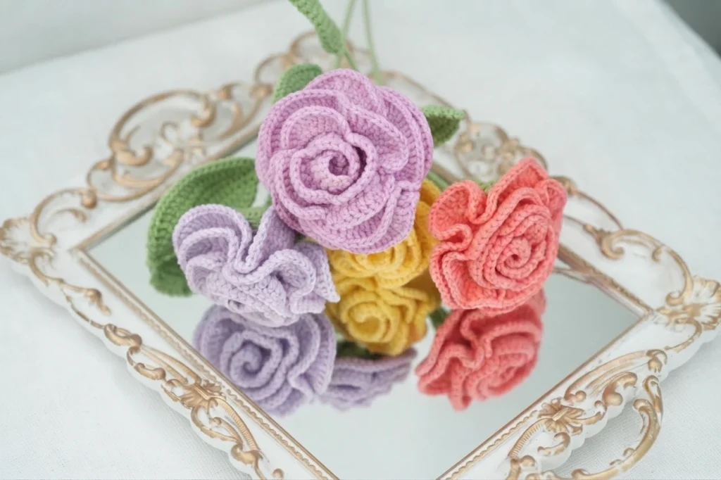Four crocheted roses in pink, lavender, yellow and coral with green leaves arranged on an ornate mirrored tray.