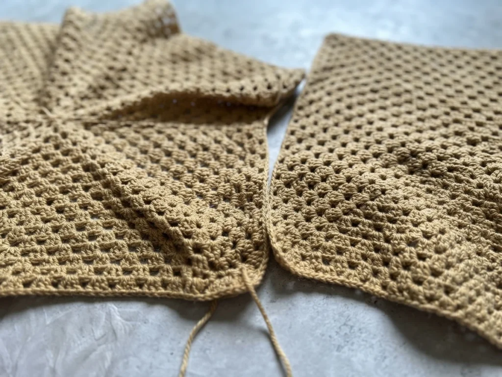 Close-up of tan granny-stitch crochet cardigan pieces laid on a pale fabric surface, with a loose yarn tail hanging down.