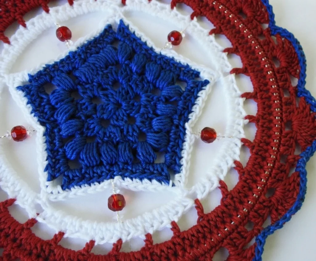 Close-up of a red, white and blue crochet mandala with a textured blue center, white edging and red faceted beads on a white background