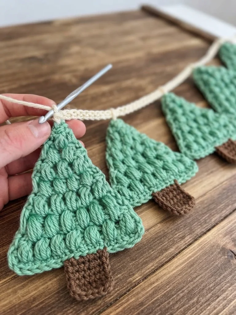 Hand holding a metal crochet hook attaching a cream crochet chain to the top of a green puff-stitch Christmas tree, with more green trees with brown trunks laid out on a wooden surface.