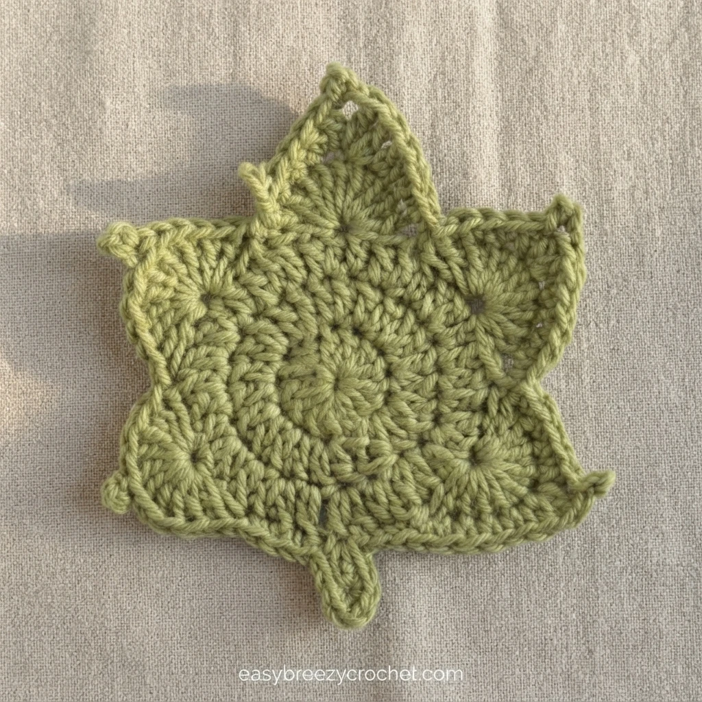 Green crocheted maple leaf shaped coaster with picot tipped points and a short stem, lying flat on light beige woven fabric, with a faint watermark near the bottom edge.