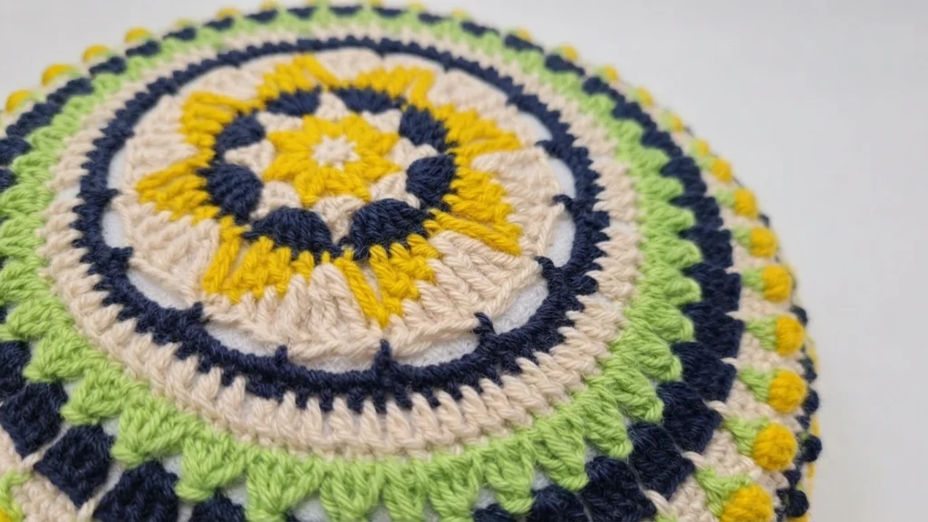 Close-up view of a round crocheted cushion with concentric mandala rings in yellow, navy blue, light green and cream around a star-shaped center.