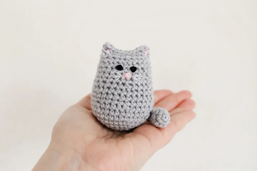 Hand holding a small grey crocheted cat with black embroidered eyes, a pink nose and white whiskers.